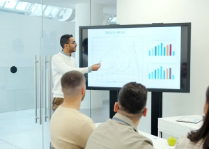 Man pointing at a line graph on a wall-mounted screen labeled Sales Value in an office
